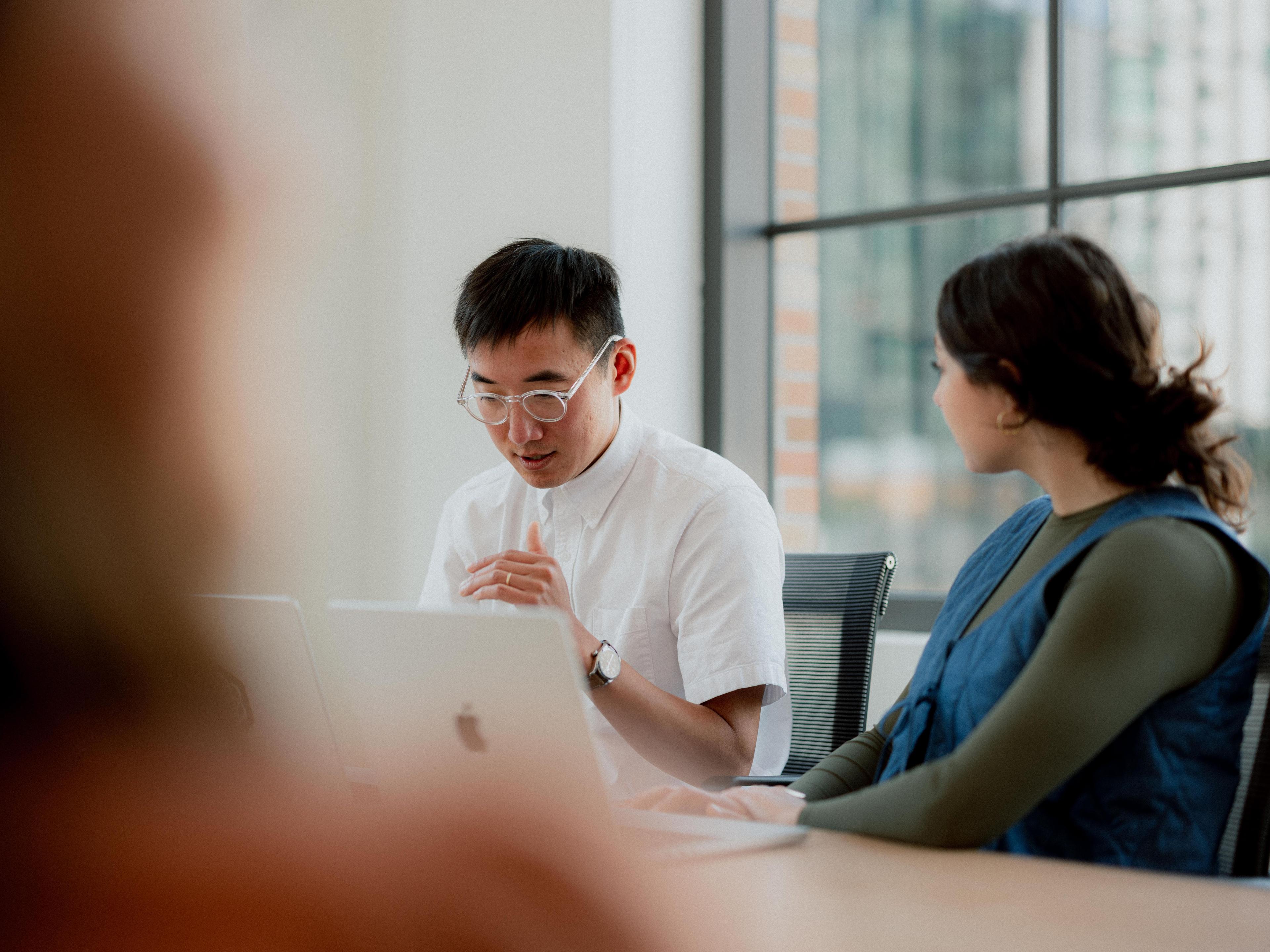 Two people sitting next to each other in a meeting. They're at a table with their laptops open, having a conversation.