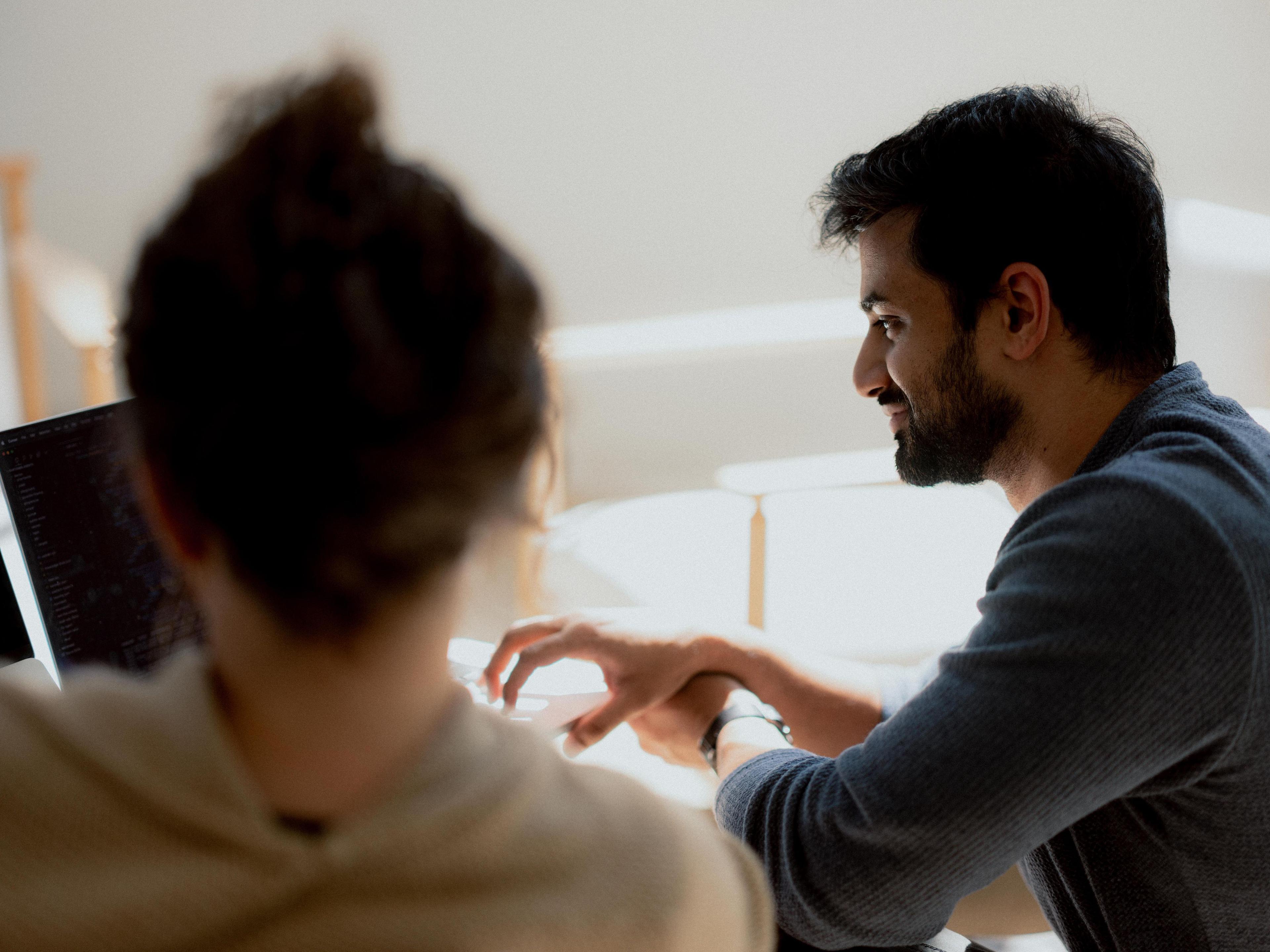A man smiles while looking at a laptop screen being used by another person.