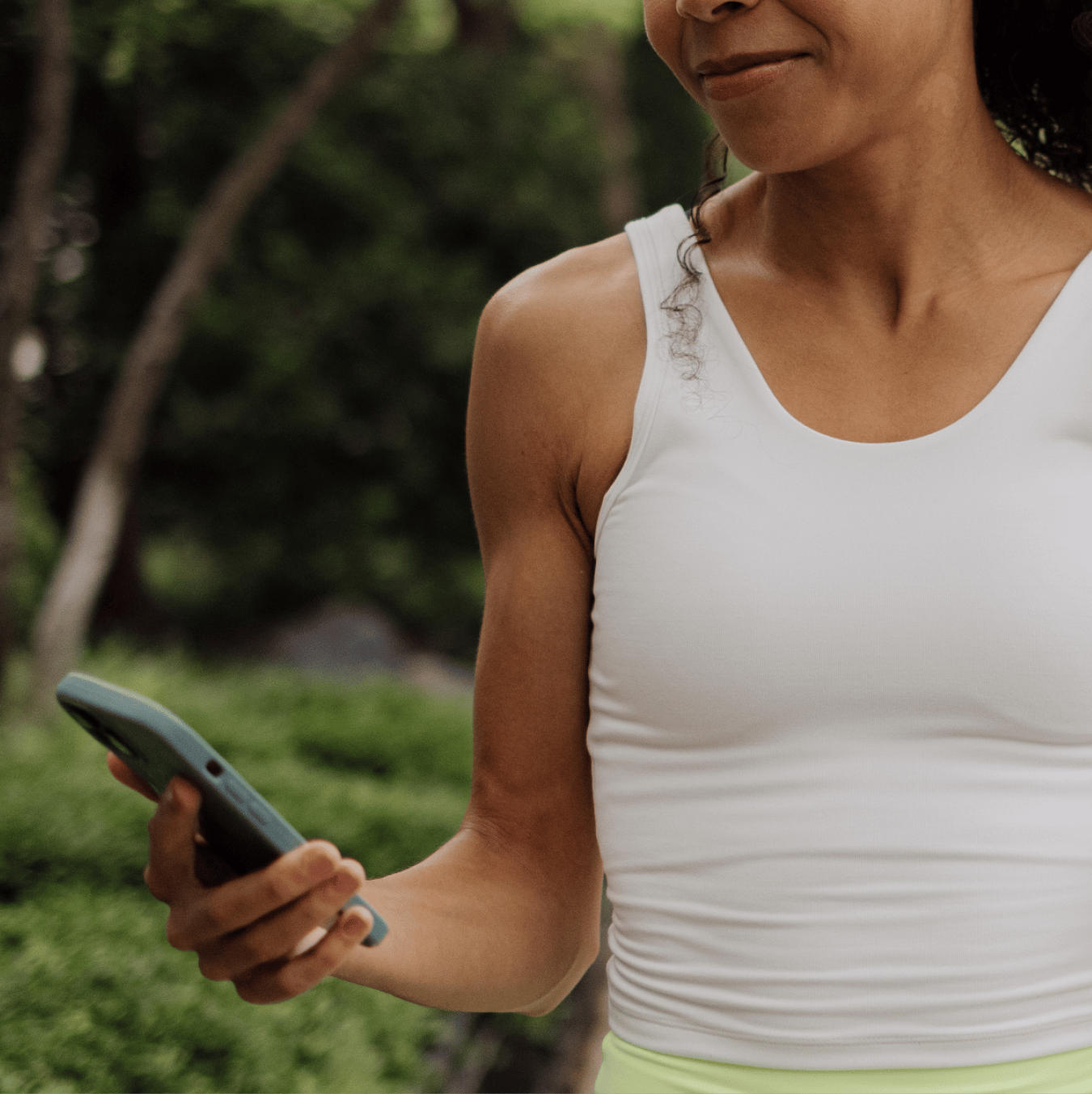 A woman in a white tank top looks at her smartphone outdoors.