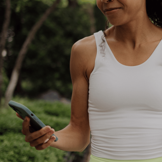 A woman in a white tank top looks at her smartphone outdoors.