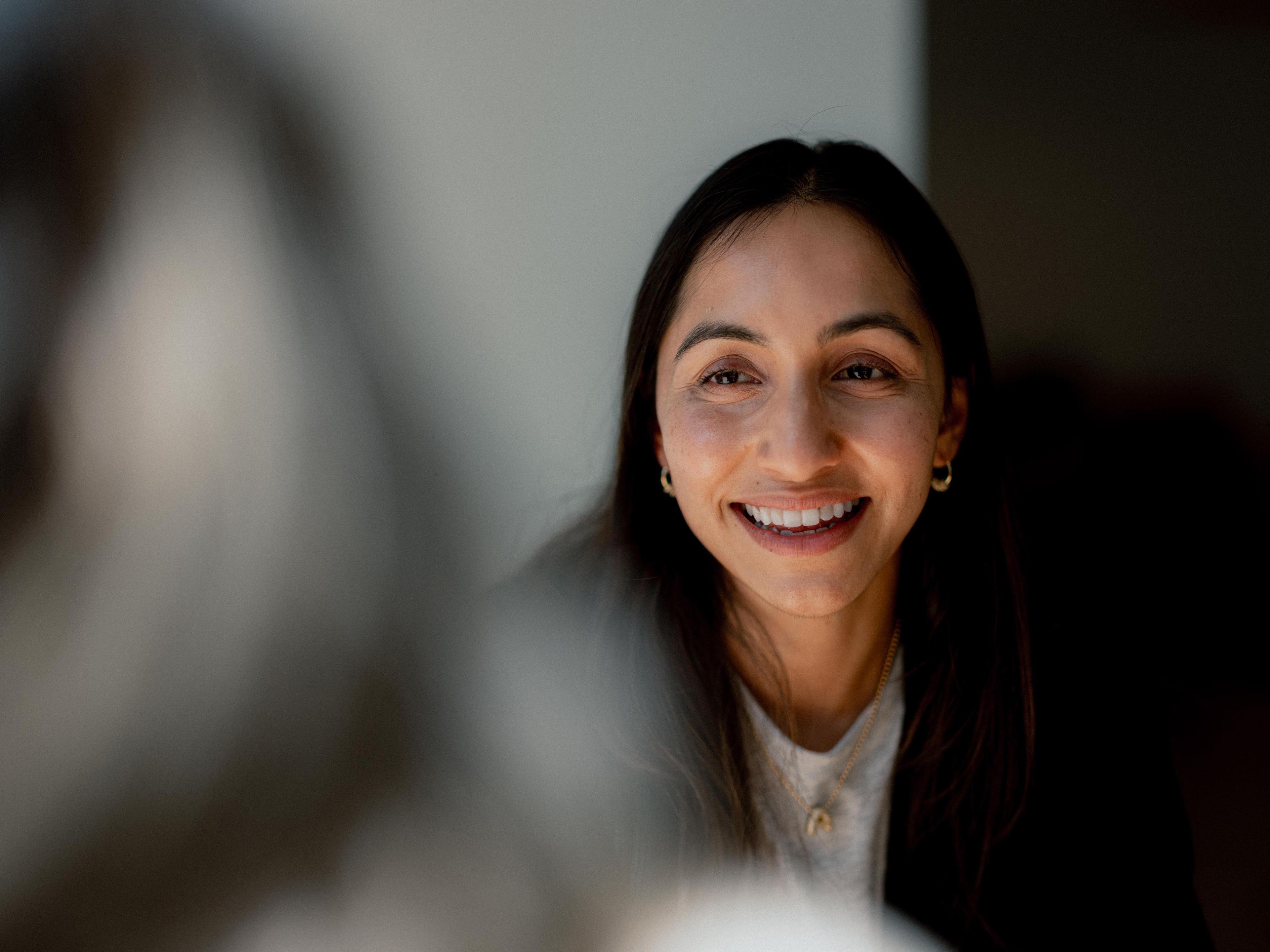 A woman smiling warmly while engaged in conversation, with soft light highlighting her face and a blurred person in the foreground.