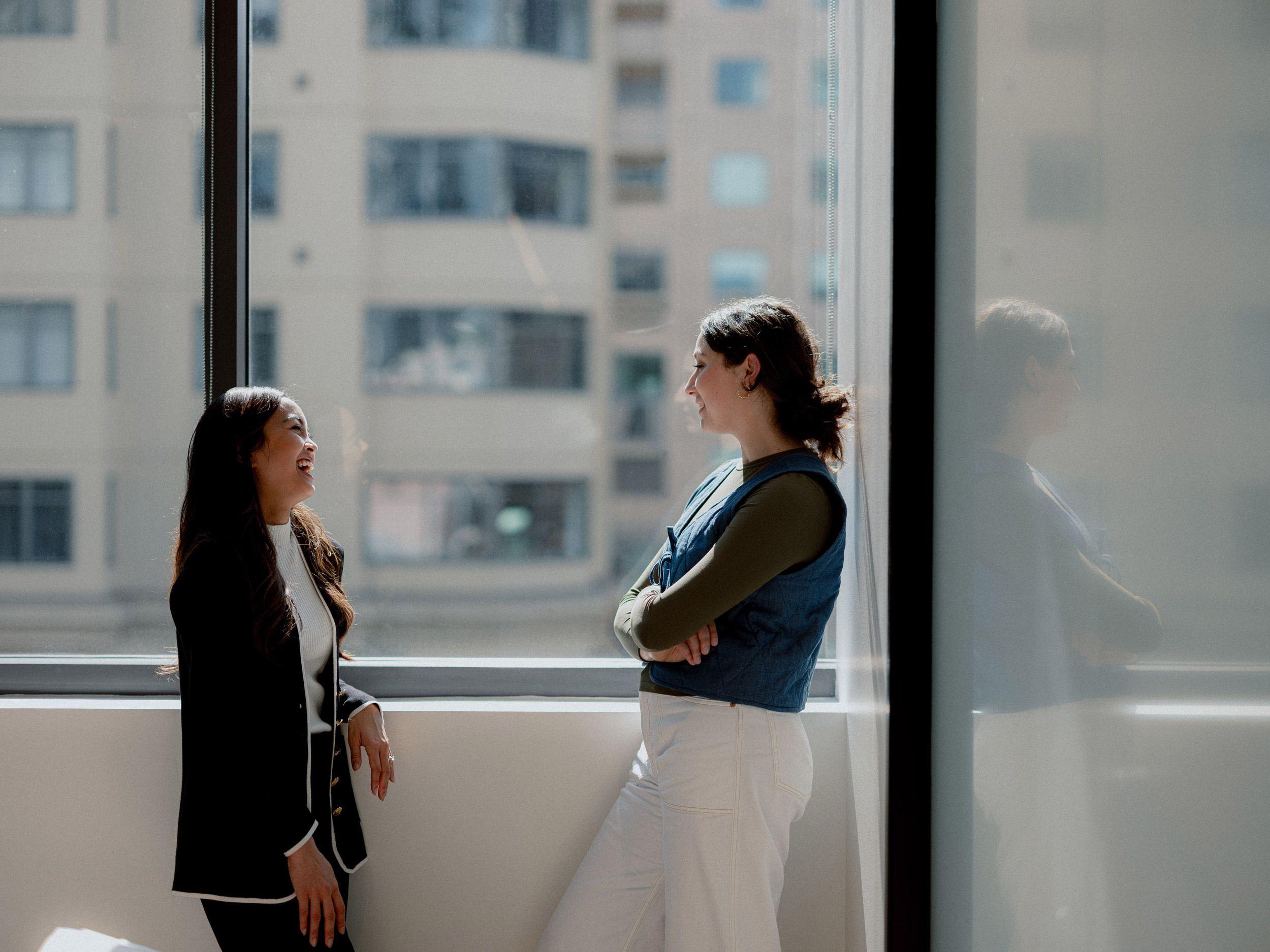 Two women standing by a large window in an office, smiling and engaged in conversation, with sunlight streaming in and buildings visible outside.