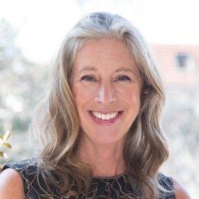 A smiling woman with long, wavy gray and blonde hair wearing a dark patterned top.